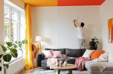 Mężczyzna maluje sufit w jasnym salonie z szarą sofą i roślinami doniczkowymi